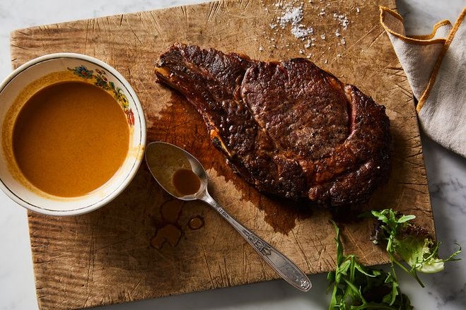 A chunk of seared steak sits on a cutting board next to a cup of orange sauce.